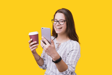 Portrait of a happy girl holding coffee cup while standing and using mobile phone on a yellow wall background