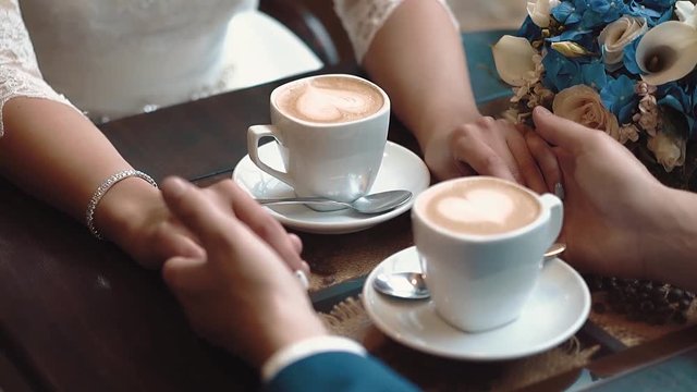 Newlyweds hold hands while sitting in a cafe, sitting at table on which there are two mugs of coffee, close-up