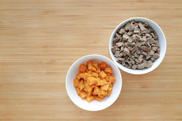Chopped of boiled pork liver and sweet potato in white bowls on wooden board. Raw healthy baby food.