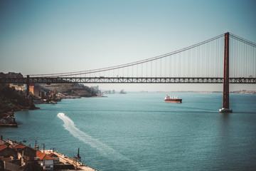 Fototapeta premium Piękne krajobrazy z ogromnym wiszącym mostem „Ponte 25 de Abril” nad rzeką Tag w Lizbonie, Portugalia w ciepły słoneczny dzień z dwoma statkami, z których jeden pozostawia długi ślad na wodzie
