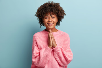 Cheerful dark skinned woman makes praying gesture, clasps palms together, smiles gladfully, wears...