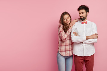 Bearded stylish adult man keeps hand crossed, feels angry wih girlfriend, wears white formal shirt with bowtie, cheerful beautiful woman leans on shoulder, looks with satisfaction at camera.