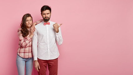 Angry bearded man looks strict and dissatisfied, points away with thumb, demonstrates something he doesnt like, puzzled woman bites lips and leans at shoulder at guy, face problem, isolated on pink