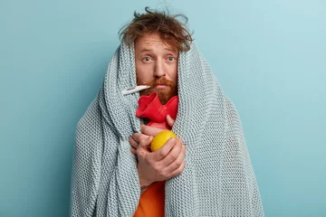Fotobehang Health care concept. Stressful sick man measures temperature, eats lemon, wrapped in plaid, looks in displeasure, cures at home, suffers from cold, fever, has symptoms of flu, isolated on blue © Wayhome Studio