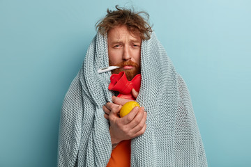 Photo of ill bearded man wrapped in coverlet, hold citrus and hot water bottle, measures temperature, trembles from cold, stay at home, has sick look, tries to break fever takes remedies. Seasonal flu - obrazy, fototapety, plakaty