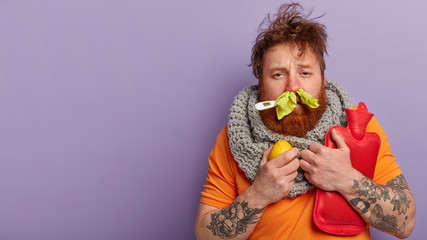 Photo of sick man has terrible sneezing, keeps napkins in nose, suffers from rhinitis, holds lemon and hot water bottle, keeps thermometer in mouth, has seasonal influenza virus, isolated on lilac - obrazy, fototapety, plakaty