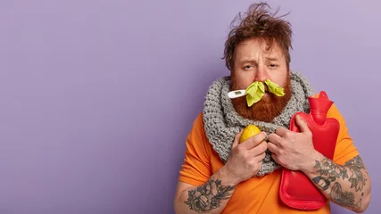 Fotobehang Photo of sick man has terrible sneezing, keeps napkins in nose, suffers from rhinitis, holds lemon and hot water bottle, keeps thermometer in mouth, has seasonal influenza virus, isolated on lilac © Wayhome Studio