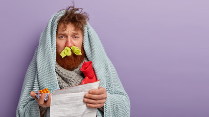 Indoor shot of displeased red haired guy holds medical leaflet with medicaments, has problems with health, suffers from running nose and flu, warms with hot water bottle and plaid, stands indoor © WHstudio Leushin N