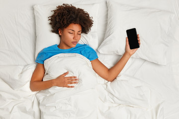 Horizontal shot of calm restful dark skinned woman falls asleep with modern smart phone, sleeps well, lies under white duvet, waits for call from boyfriend. Sleeping, technology, people, rest concept © WHstudio Leushin N