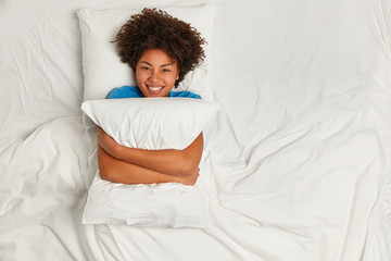 Playful pleased dark skinned woman embraces pillow, looks gladfully at camera, smiles broadly,...