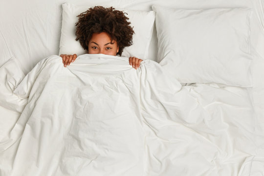 Cheerful black woman hides under soft white blanket, has fun in bed, rejoices good morning, has curly hair, rests in bed. View from above. Copy space. People, ethnicity and sleeping concept.
