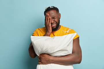 Dejected crying man covers face with hand, has thick stubble, dressed in yellow t shirt, has problems with sleep, carries white pillow, isolated over blue background. Sleeping and depression