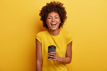 Monchrome shot of cheerful dark skinned woman laughs and talks casually, holds disposable cup of...
