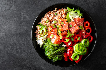 poke bowl of brown rice, trout or salmon fillet, Chuka Seaweed Salad, sesame and fresh vegetables on dark background, top view, copy space, healthy food trend