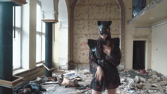 Young beautiful girl stands on drum among the garbage in ruined building. She wears leather sword, and cat's mask made of leather. She dances, depicting cat, close-up