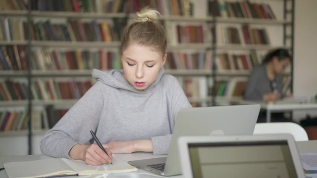 Pan shot of young blonde female preparing for exams in library. Female student writing in the notebook sitting in the library. Background of long bookshelf in the library.