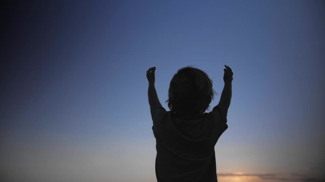 Child silhouette raising hands up in the air on blue sky background. Praying, enjoying nature outdoors in summer sunset