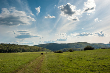 Hikers on green meadow with dramatic cloudy sky. Back view of group of people hiking in low mountains. Hungarian Karst, Aggtelek Karst, Europe.