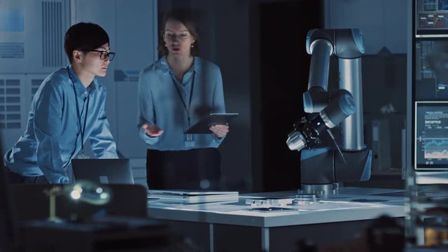 Man and Woman Engineers with Laptop and a Tablet Analyse and Discuss How a Futuristic Robotic Arm Works and Moves a Metal Object. They are in a High Tech Research Laboratory with Modern Equipment.