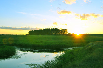 sunset landscape with mirror river and blue sky in green field.