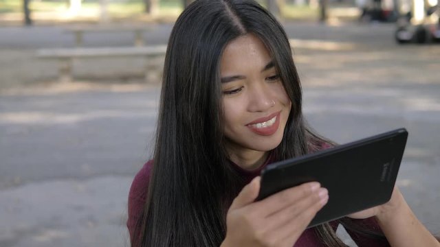 Asian attractive woman scrolling digital tablet in the park and smiling