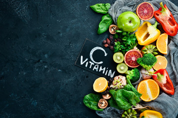 Fruits and vegetables that contain vitamin C: Orange, lemon, apple, roses, garlic, broccoli, apple, kiwi, spinach. Top view. On a black stone background. - obrazy, fototapety, plakaty