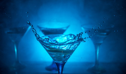 Martini cocktail glass in hand splashing on dark toned smoky background or colorful cocktail in glass with splashes and olives. Party club entertainment. Mixed light. Selective focus