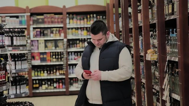 Fat man in the supermarket is in the department with alcohol and looks into the phone.