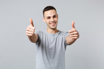 Portrait of smiling handsome young man in casual clothes standing, looking camera, showing thumbs up isolated on grey wall background. People sincere emotions, lifestyle concept. Mock up copy space.