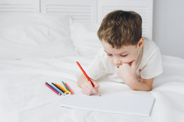 Boy draws with colorful pencils sitting on the bed