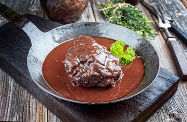 Traditional German braised beef cheeks in brown red wine sauce with herbs as closeup in a wrought-iron skillet on an old burnt board