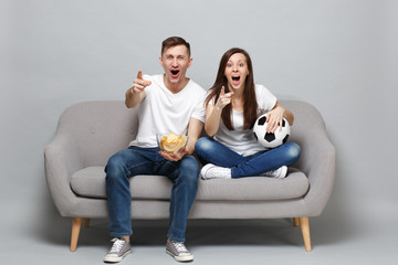 Fun couple woman man football fans cheer up support favorite team with soccer ball hold glass bowl of chips, pointing index fingers on camera isolated on grey background. Sport family leisure concept.