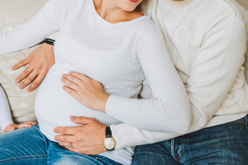 Husband and pregnant wife with their hands on the belly. Couple wearing white sweaters and jeans. Concept of pregnancy and expecting a baby.