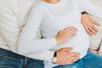 Husband and pregnant wife with their hands on the belly. Couple wearing white sweaters and jeans. Concept of pregnancy and expecting a baby.
