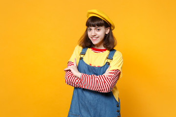 Portrait of smiling girl teenager in french beret and denim sundress holding hands crossed isolated on yellow wall background in studio. People sincere emotions, lifestyle concept. Mock up copy space.