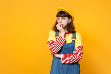 Portrait of nervous girl teenager in french beret, denim sundress gnawing nails, looking aside isolated on yellow background in studio. People sincere emotions lifestyle concept. Mock up copy space.