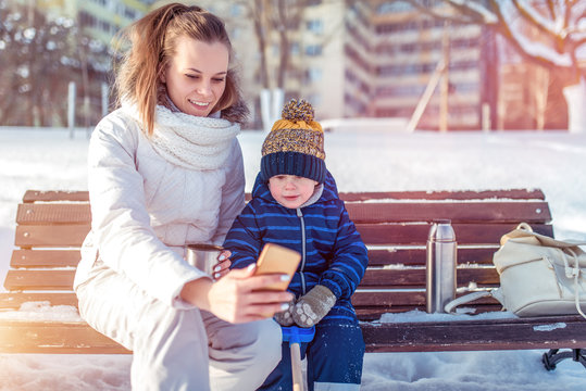Mom takes a picture and shows smartphone to her child a little boy. In winter in the city on a bench, fresh frosty air in December and January.