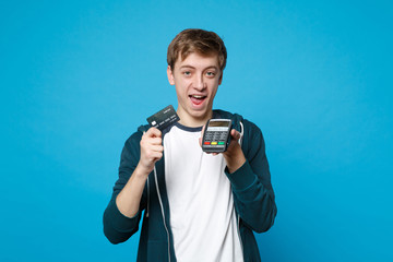 Cheerful young man holding wireless modern bank payment terminal to process and acquire credit card payments isolated on blue background. People sincere emotions lifestyle concept. Mock up copy space.
