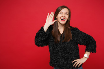 Cheerful curious young woman in black fur sweater eavesdrop with hearing gesture near ear isolated...