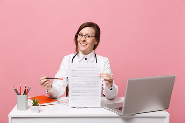 Beautiful female doctor sits at desk works on computer with medical document in hospital isolated on pastel pink wall background. Woman in medical gown glasses stethoscope. Healthcare medicine concept