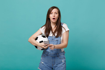Shocked concerned young girl football fan holding soccer ball using mobile phone isolated on blue...