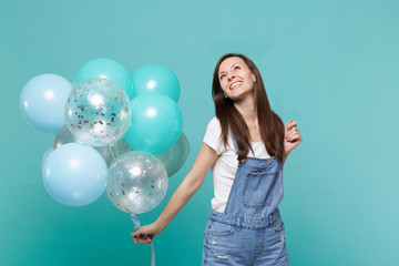 Smiling happy pensive young woman in denim clothes looking up celebrating and holding colorful air balloons isolated on blue turquoise wall background. Birthday holiday party, people emotions concept.