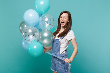 Funny young woman in denim clothes blinking, keeping mouth wide open, celebrating, holding colorful air balloons isolated on blue turquoise background. Birthday holiday party, people emotions concept.