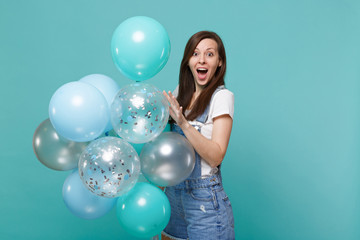 Excited young woman in denim clothes keeping mouth wide open, celebrating and holding colorful air...