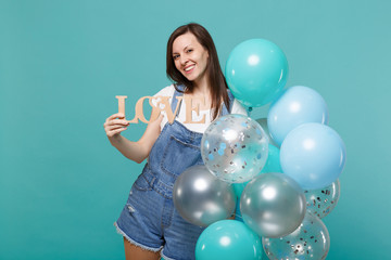 Fototapeta premium Smiling young woman in denim clothes holding wooden word letters love, celebrating with colorful air balloons isolated on blue turquoise background. Birthday holiday party, people emotions concept.