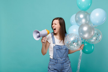 Irritated young woman looking aside screaming in megaphone while celebrating, holding colorful air balloons isolated on blue turquoise wall background. Birthday holiday party, people emotions concept.