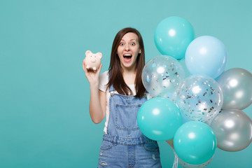 Fototapeta premium Surprised young woman keeping mouth wide open, holding piggy money bank, celebrating with colorful air balloons isolated on blue turquoise background. Birthday holiday party, people emotions concept.