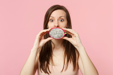 Shocked young woman covering mouth with half of fresh ripe pitahaya, dragon fruit isolated on pink pastel wall background in studio. People vivid lifestyle relax vacation concept. Mock up copy space.