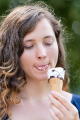 Woman eating one vanilla chocolate ice cream gelato cone looking down tongue licking with bokeh background of park in Europe city during sunny summer day
