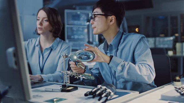 Japanese Development Engineer in Blue Shirt is Soldering a Circuit Board in a High Tech Research Laboratory with Modern Equipment. His Colleague Asks Him a Questio and Points on the Compter Screen.